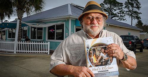 Ned Pollack wearing a hat stands outside a light teal building, cradling a cookbook titled "Ned's Kitchen." He's ready to launch his campaign to inspire others with his love for fresh produce.