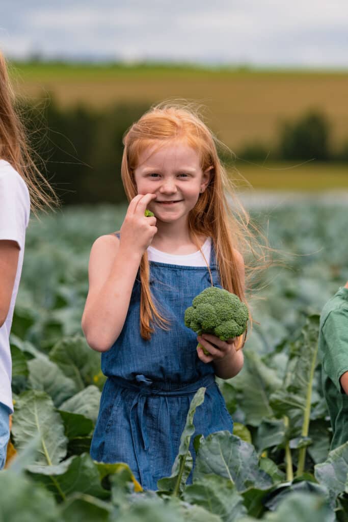 A young girl with red hair stands in a broccoli field at Smith’s Farm, holding a piece of broccoli and smiling for what could easily be one of her first professional headshots. She's wearing a blue dress, ready to charm the Global Produce & Floral Show with her bright smile.