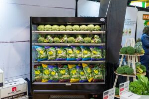 A refrigerated display of broccoli heads on the top shelf and various packaged leafy greens on the middle and bottom shelves.