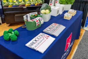 Display table featuring broccoli and cauliflower merchandise, promotional flyers, and branded materials, including shirts and brochures.