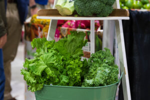 A variety of leafy greens, including lettuce, kale, and broccoli, are displayed in a metal basket and on a shelf at a market.