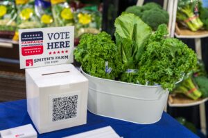 A display of fresh kale in a white container with a sign that reads "Vote Here" at a market stand, next to a white ballot box.