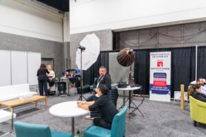 People setting up a photography station at a trade show booth, with lights and banners, surrounded by tables and chairs.